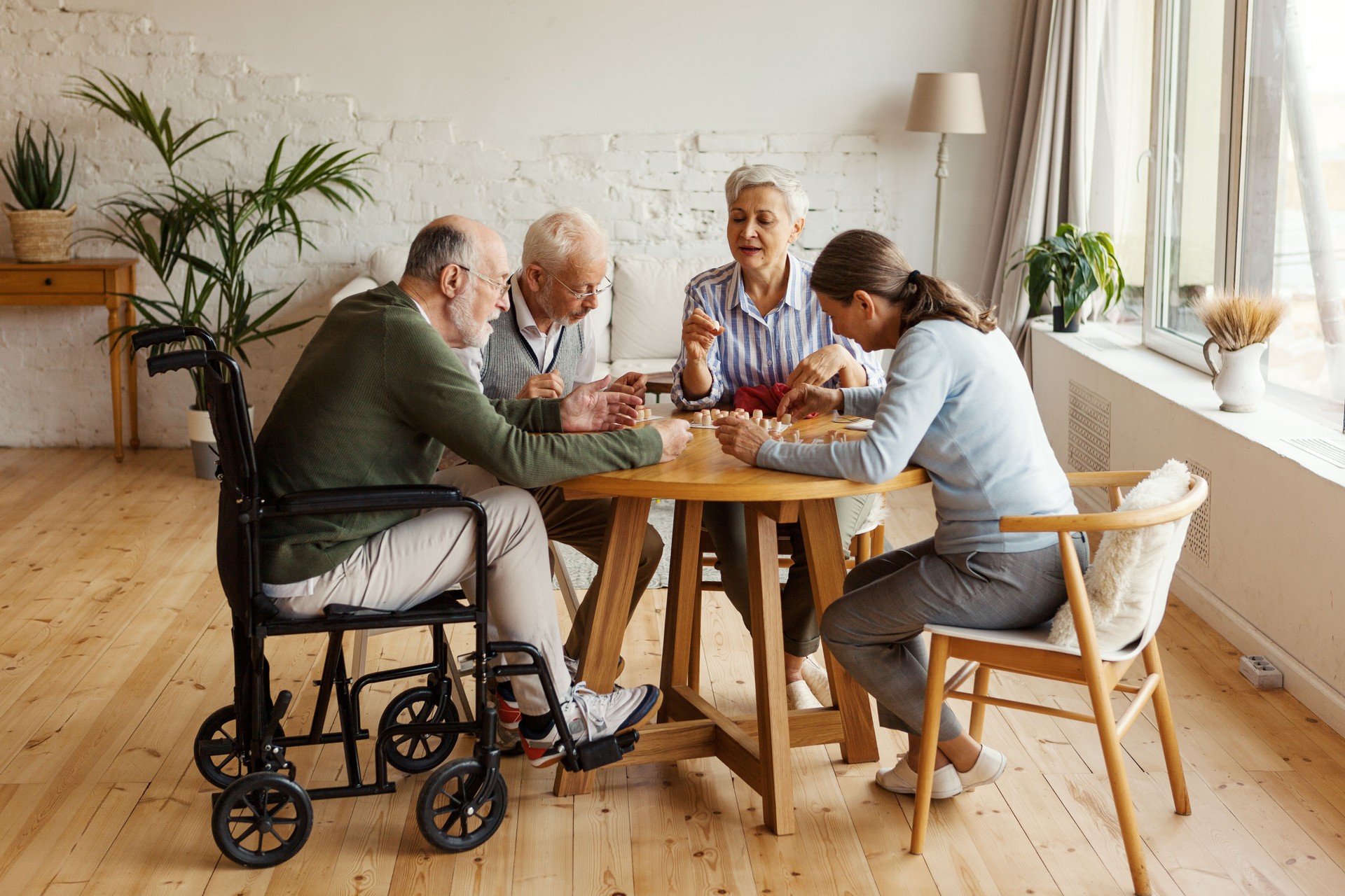 Group of four senior people, two men including disabled one and two women, sitting at table together and playing bingo game in nursing home
